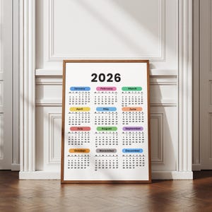 May include: A framed 2026 calendar with a wooden frame. The calendar displays the months of the year in a grid format, with each month's name in a different color. The year "2026" is printed in large, bold letters at the top.