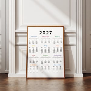 May include: A vertical wall calendar for the year 2027 featuring a white background with black text for the months and dates. Each month name is displayed in a different pastel color. The calendar is enclosed in a simple brown wooden frame.
