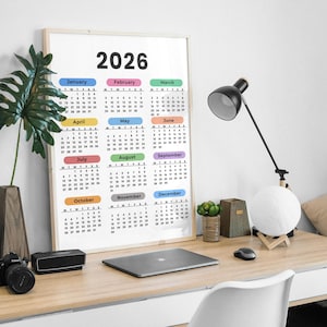 May include: A framed 2026 calendar with a white background and colorful month labels. The calendar is on a wooden desk with a black desk lamp, a laptop, and a camera.
