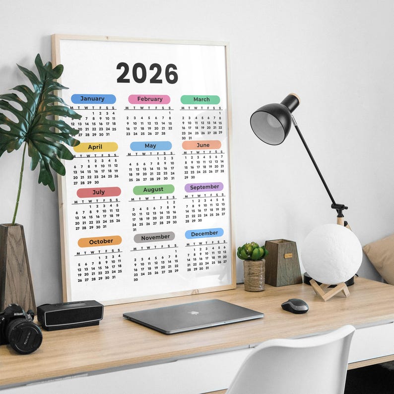 May include: A framed 2026 calendar with a white background and colorful month labels. The calendar is on a wooden desk with a black desk lamp, a laptop, and a camera.
