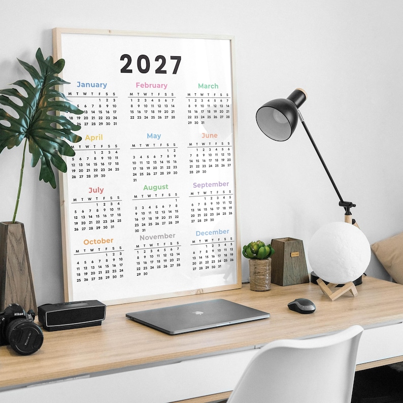 May include: A vertical wall calendar for the year 2027 featuring a minimalist white background with black text for the months and dates. Each month name is displayed in a different pastel color, organized in a grid layout with twelve sections.