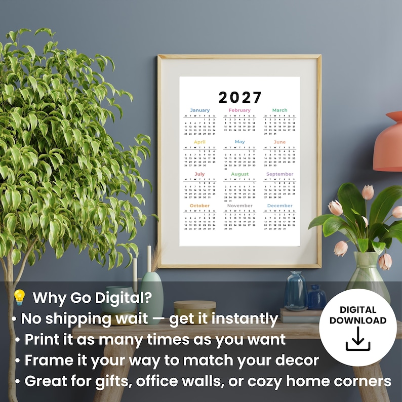 May include: A framed 2027 wall calendar print featuring a white background with black text for the months and dates. Each month name is displayed in a different color. The calendar is presented in a light wood frame hanging on a wall.