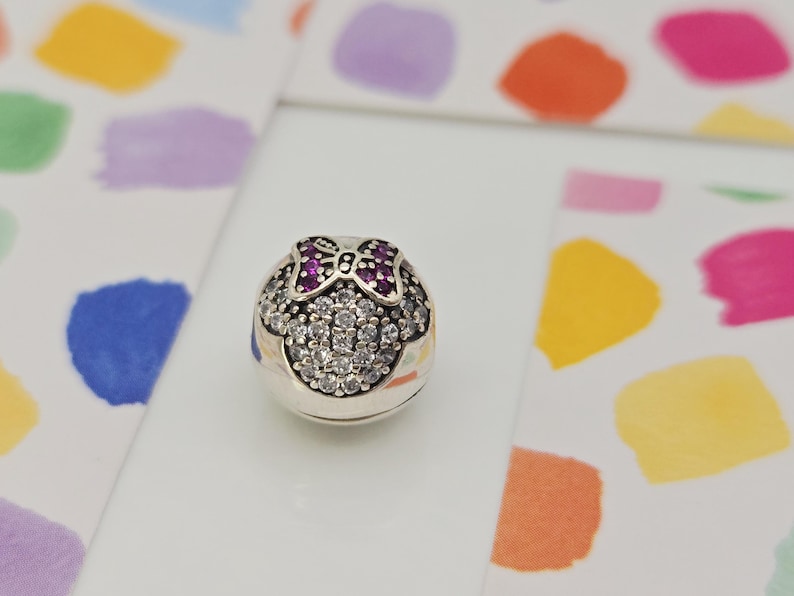 May include: A silver charm with a Minnie Mouse design. The charm features a bow with pink gemstones and a face made of clear gemstones. The charm is on a white surface with colorful paint splotches in the background.
