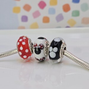 Puede incluir: Tres cuentas decorativas en una cadena de plata. Las cuentas presentan lunares rojos, un dise&ntilde;o de Minnie Mouse y negro con huellas de patas blancas. Las cuentas son probablemente para una pulsera de abalorios.