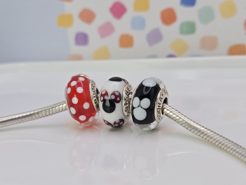 Puede incluir: Tres cuentas decorativas en una cadena de plata. Las cuentas presentan lunares rojos, un dise&ntilde;o de Minnie Mouse y negro con huellas de patas blancas. Las cuentas son probablemente para una pulsera de abalorios.