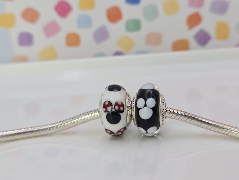 May include: Two decorative beads on a silver chain. One bead is white with a black circle and red polka dots. The other bead is black with white paw prints. The beads are likely for a bracelet or necklace.