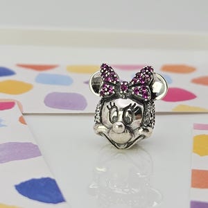 May include: A silver charm depicting Minnie Mouse's face. The charm features a bow adorned with purple gemstones and sparkling details. The background is a white surface with colourful paint splotches.