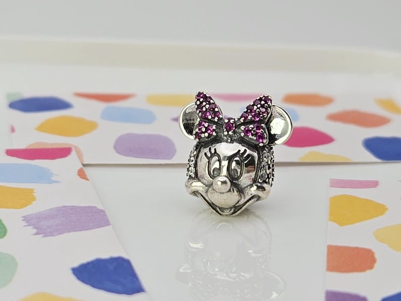 May include: A silver charm depicting Minnie Mouse's face. The charm features a bow adorned with purple gemstones and sparkling details. The background is a white surface with colourful paint splotches.