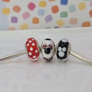 Puede incluir: Tres abalorios decorativos en una pulsera de plata. Las cuentas son rojas con lunares blancos, blancas con un dise&ntilde;o de Minnie Mouse y negras con huellas de patas blancas. El fondo es un patr&oacute;n colorido.