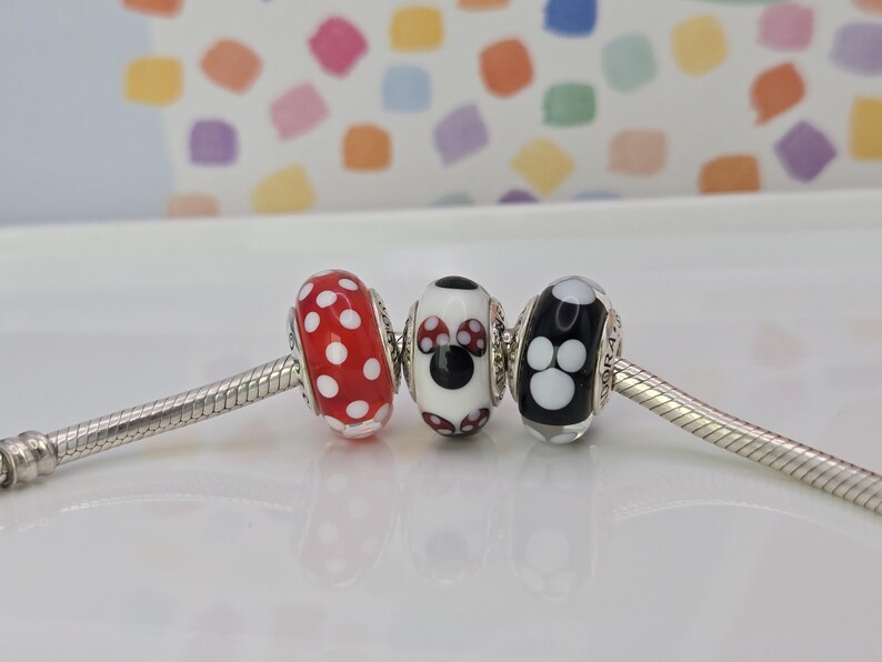 Puede incluir: Tres abalorios decorativos en una pulsera de plata. Las cuentas son rojas con lunares blancos, blancas con un dise&ntilde;o de Minnie Mouse y negras con huellas de patas blancas. El fondo es un patr&oacute;n colorido.