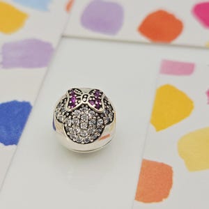 May include: A silver charm with a Minnie Mouse design. The charm features a bow with purple gems and a face outlined with clear crystals. The charm is round and sits on a white surface with colorful paint splotches in the background.
