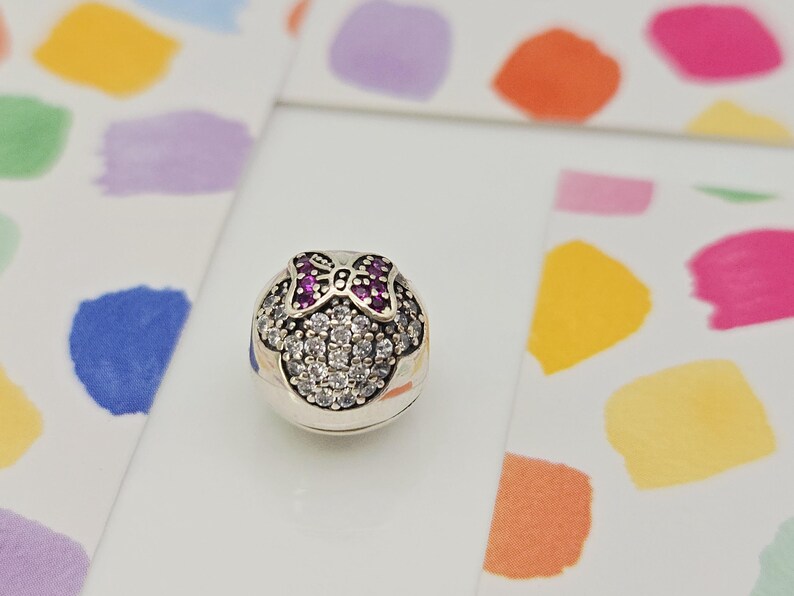 May include: A silver charm with a Minnie Mouse design. The charm features a bow with purple gems and a face outlined with clear crystals. The charm is round and sits on a white surface with colorful paint splotches in the background.
