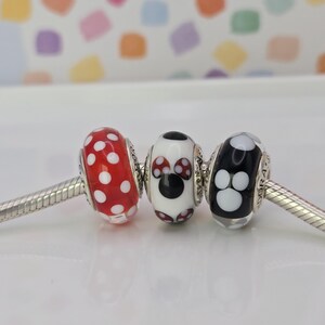 Puede incluir: Tres cuentas decorativas en una cadena plateada. La primera cuenta es roja con lunares blancos. La segunda cuenta es blanca con un dise&ntilde;o de Mickey Mouse negro. La tercera cuenta es negra con huellas de patas blancas.