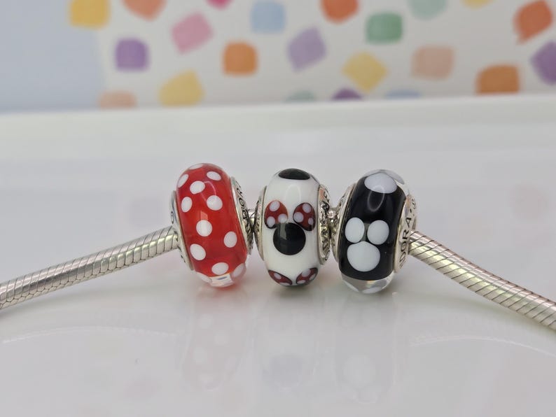 Puede incluir: Tres cuentas decorativas en una cadena plateada. La primera cuenta es roja con lunares blancos. La segunda cuenta es blanca con un dise&ntilde;o de Mickey Mouse negro. La tercera cuenta es negra con huellas de patas blancas.