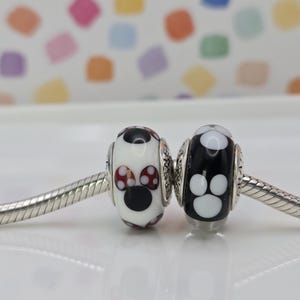 May include: Two decorative beads on a silver chain. One bead is white with a black face and red polka dot accents. The other bead is black with white paw prints. The beads are likely for a bracelet or necklace.