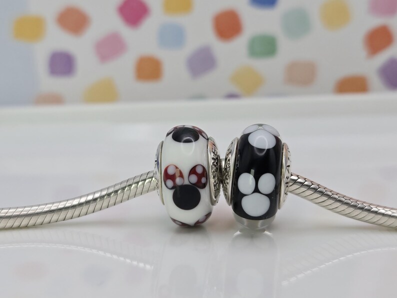 May include: Two decorative beads on a silver chain. One bead is white with a black face and red polka dot accents. The other bead is black with white paw prints. The beads are likely for a bracelet or necklace.