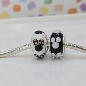 May include: Two decorative beads with a silver core. One bead is white with a black Mickey Mouse design and red accents. The other bead is black with a white paw print design. The beads are on a silver chain.