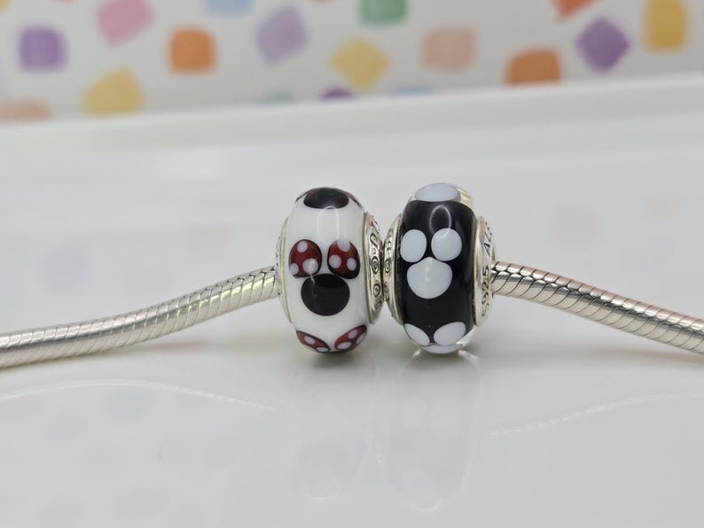 May include: Two decorative beads with a silver core. One bead is white with a black Mickey Mouse design and red accents. The other bead is black with a white paw print design. The beads are on a silver chain.