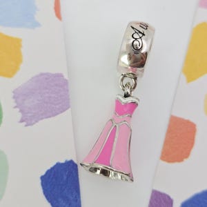 May include: A silver charm with the word "Au" in cursive script. The charm features a pink and silver dress pendant. The dress has a silver outline and is filled with shades of pink. The charm is designed to be attached to a bracelet or necklace.