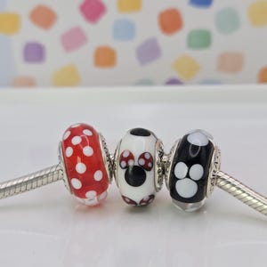 Puede incluir: Tres cuentas decorativas en una cadena plateada. Las cuentas son rojas con lunares blancos, blancas con un dise&ntilde;o de Minnie Mouse y negras con huellas de patas blancas. El fondo es un patr&oacute;n colorido.