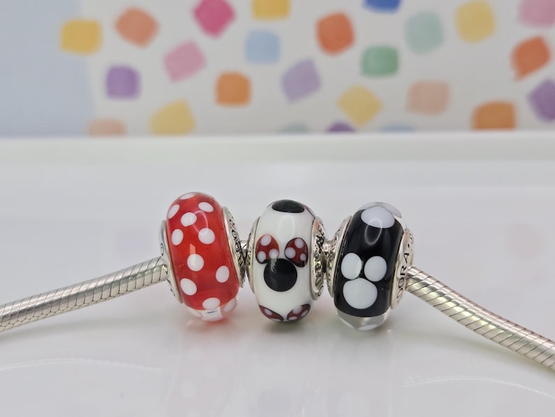 Puede incluir: Tres cuentas decorativas en una cadena plateada. Las cuentas son rojas con lunares blancos, blancas con un dise&ntilde;o de Minnie Mouse y negras con huellas de patas blancas. El fondo es un patr&oacute;n colorido.