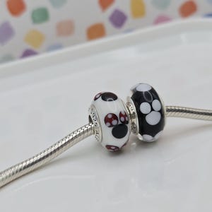 May include: Two glass beads with a paw print design on a silver chain. One bead is white with black and red paw prints, and the other is black with white paw prints. The beads are likely for a bracelet or necklace.