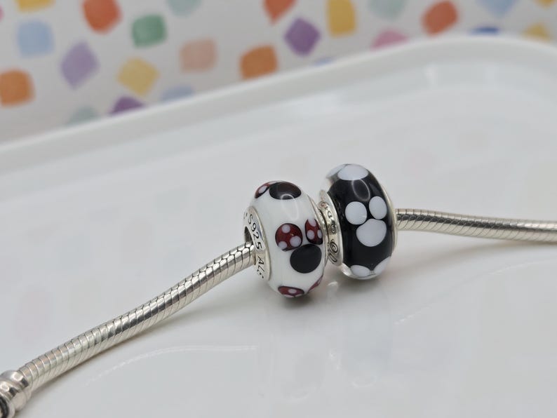 May include: Two glass beads with a paw print design on a silver chain. One bead is white with black and red paw prints, and the other is black with white paw prints. The beads are likely for a bracelet or necklace.