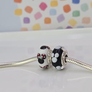 May include: Two decorative beads on a silver chain. One bead is white with a black and red design. The other bead is black with white paw prints. The beads are round and have silver accents. The background is a white surface with a colourful pattern.