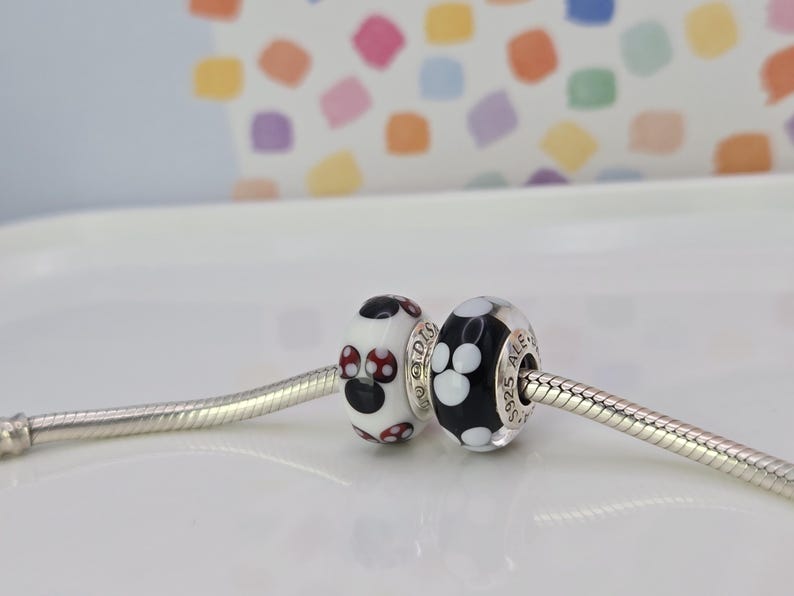 May include: Two decorative beads on a silver chain. One bead is white with a black and red design. The other bead is black with white paw prints. The beads are round and have silver accents. The background is a white surface with a colourful pattern.