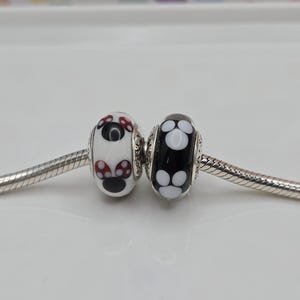 May include: Two glass beads with silver detailing. One bead is white with a red and black design, the other is black with a white design. Both beads are on a silver chain, suitable for a charm bracelet.
