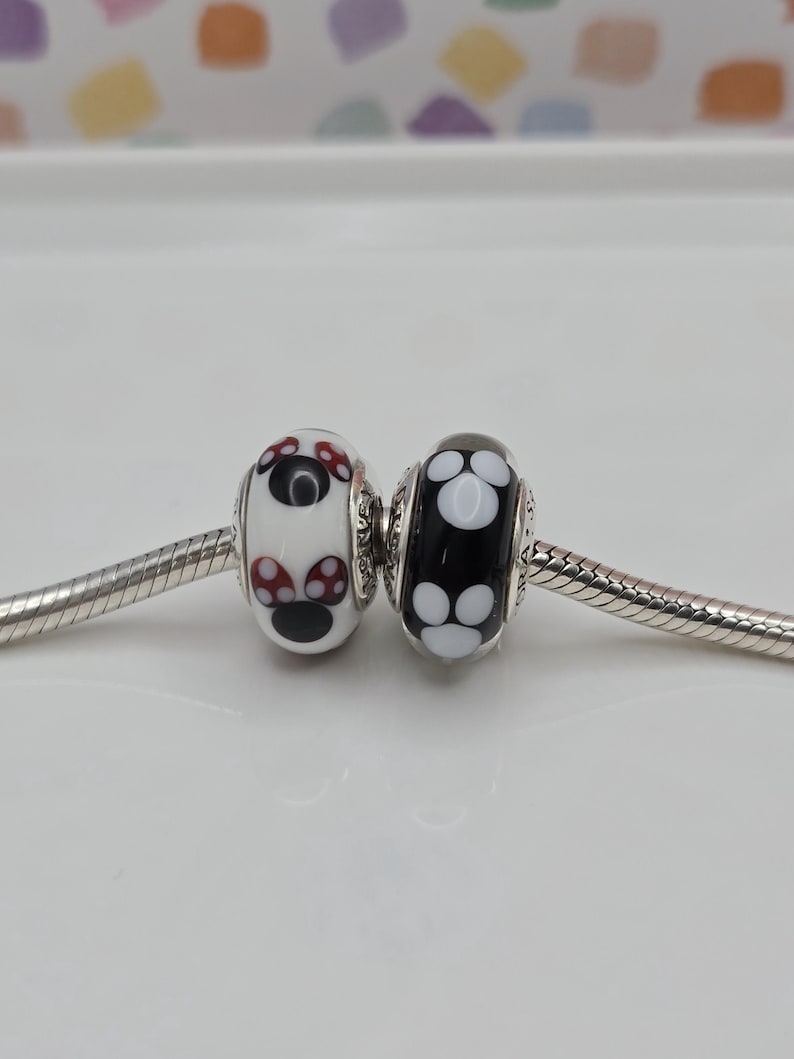 May include: Two glass beads with silver detailing. One bead is white with a red and black design, the other is black with a white design. Both beads are on a silver chain, suitable for a charm bracelet.