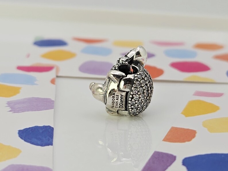 May include: A silver charm with a rounded, textured body encrusted with small, clear crystals. The charm has a small, smooth silver head and a clasp. The charm is stamped with "PANDORA S925 ALE &copy;Disney".