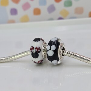 May include: Two decorative beads on a silver chain. One bead is white with black and red polka dots, the other is black with white paw prints. The beads are likely for a bracelet or necklace.