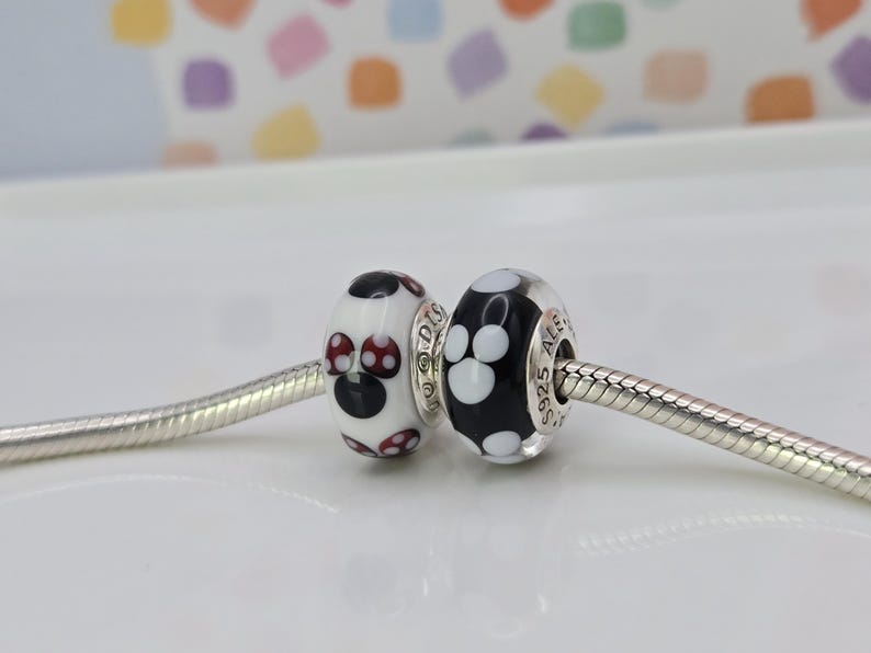 May include: Two decorative beads on a silver chain. One bead is white with black and red polka dots, the other is black with white paw prints. The beads are likely for a bracelet or necklace.