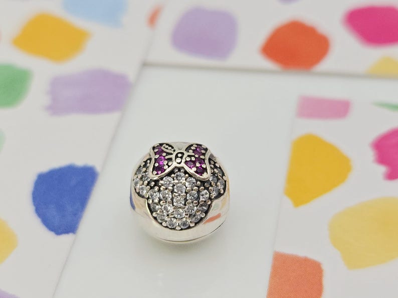 May include: A silver charm with a Minnie Mouse design. The charm features a bow with purple stones and a face outlined with clear stones. The charm is on a white surface with colorful abstract shapes in the background.