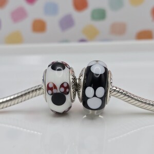 May include: Two glass beads on a silver chain. One bead is white with a black nose and red and white polka dot ears. The other bead is black with white paw prints.