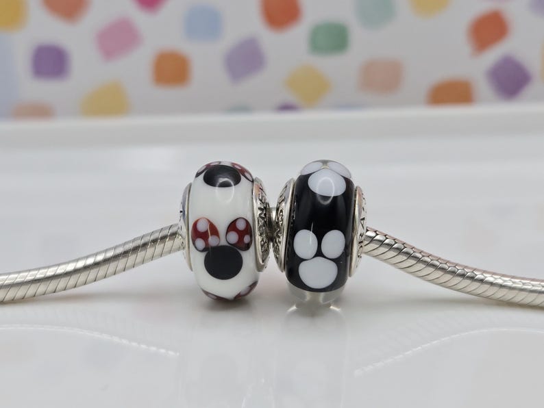 May include: Two glass beads on a silver chain. One bead is white with a black nose and red and white polka dot ears. The other bead is black with white paw prints.