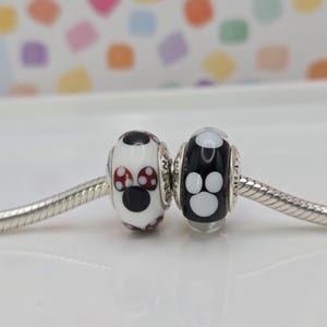 May include: Two decorative beads on a silver chain. One bead is white with black and red mushroom-like designs. The other bead is black with white paw print designs. Jewellery.