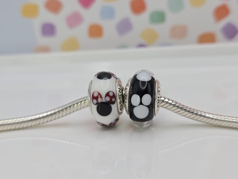 May include: Two decorative beads on a silver chain. One bead is white with black and red mushroom-like designs. The other bead is black with white paw print designs. Jewellery.
