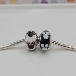 May include: Two decorative glass beads. One is white with red and black mushroom designs, the other is black with white flower patterns. Both are strung on a silver chain.