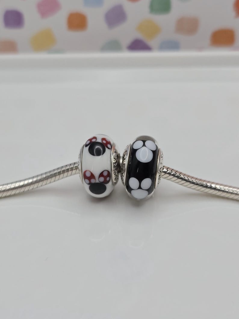 May include: Two decorative glass beads. One is white with red and black mushroom designs, the other is black with white flower patterns. Both are strung on a silver chain.