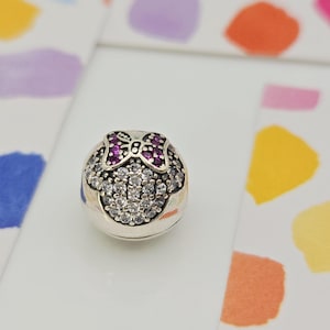 May include: A silver charm with a Minnie Mouse design. The charm features a bow with purple gems and a face outlined with clear gems. The charm is on a white surface with colorful paint splotches in the background.