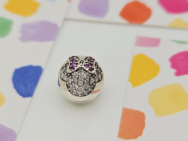 May include: A silver charm with a Minnie Mouse design. The charm features a bow with purple gems and a face outlined with clear gems. The charm is on a white surface with colorful paint splotches in the background.