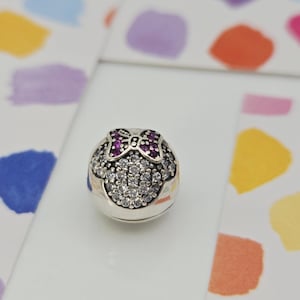 May include: A silver charm with a Minnie Mouse design. The charm features a bow with pink gemstones and a face outlined with clear gemstones. The charm is round and has a polished finish. The background has colorful abstract shapes.