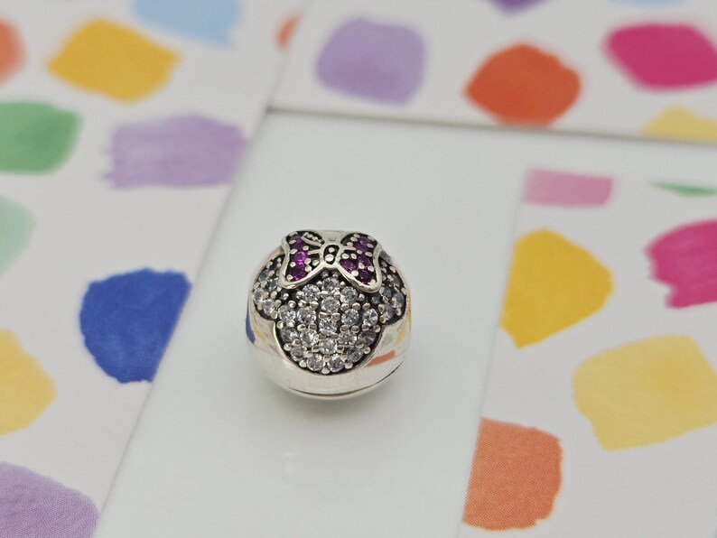 May include: A silver charm with a Minnie Mouse design. The charm features a bow with pink gemstones and a face outlined with clear gemstones. The charm is round and has a polished finish. The background has colorful abstract shapes.