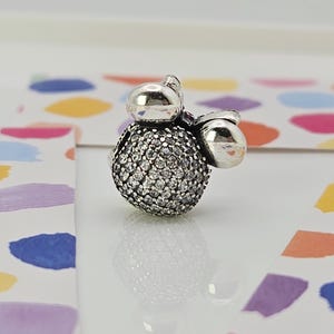 May include: A silver charm with a round, gem-studded centre and two smaller, smooth silver spheres. The charm is likely a bead or pendant, designed for a bracelet or necklace. The background features colourful abstract shapes.