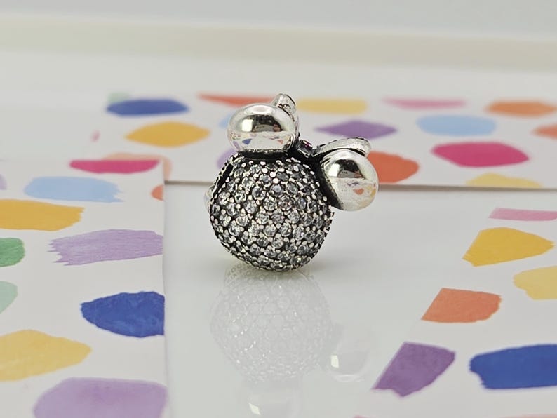 May include: A silver charm with a round, gem-studded centre and two smaller, smooth silver spheres. The charm is likely a bead or pendant, designed for a bracelet or necklace. The background features colourful abstract shapes.