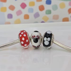 Puede incluir: Tres cuentas decorativas en una pulsera de plata. Las cuentas son rojas con lunares blancos, un dise&ntilde;o de Minnie Mouse y una cuenta negra con huellas de patas blancas. El fondo es un patr&oacute;n colorido.