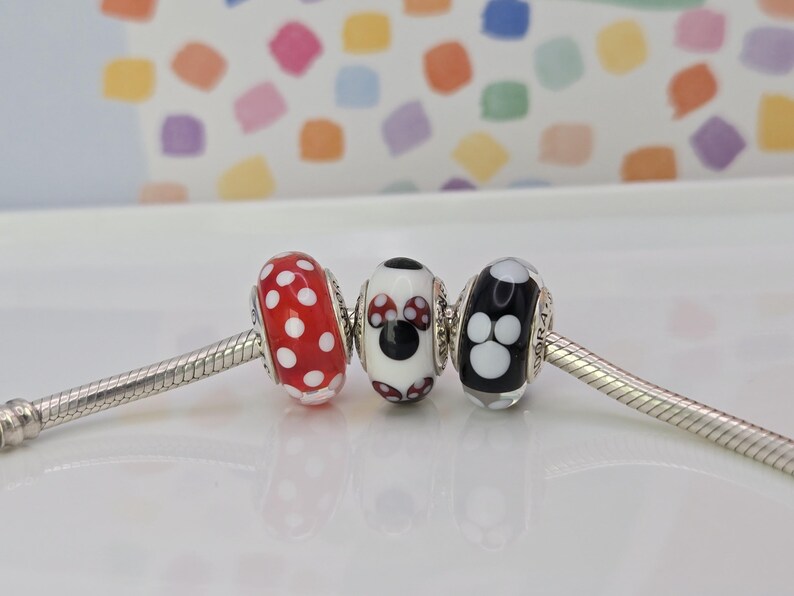 Puede incluir: Tres cuentas decorativas en una pulsera de plata. Las cuentas son rojas con lunares blancos, un dise&ntilde;o de Minnie Mouse y una cuenta negra con huellas de patas blancas. El fondo es un patr&oacute;n colorido.