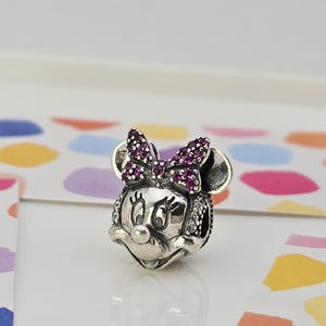 May include: A silver Minnie Mouse charm with a bow embellished with small, dark pink stones. The charm has a detailed design with small, clear stones around the face and ears. The charm is on a white surface with colourful shapes.